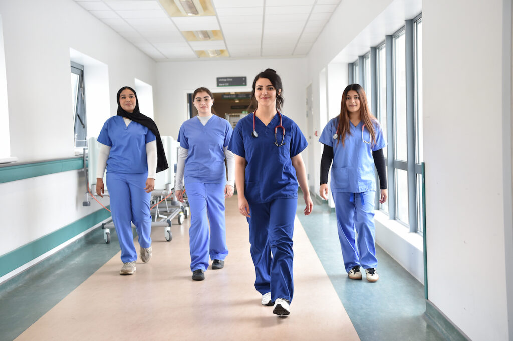 Dr. Ayah Albani, Aliyah Bangi, Safiyya Hanslod and Azin Ismael walking together in blue scrubs along a hospital corridor.